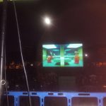 Vive la experiencia única de cine al aire libre con una proyección animada al estilo LEGO, presentada por MovieMovil la empresa del cine al aire libre en un entorno nocturno inspirador. Cine al aire libre en ambiente nocturno con pantalla que proyecta una escena animada de personajes al estilo LEGO, acompañada de una barrera azul y un poste negro, organizada por MovieMovil la empresa del cine al aire libre.
