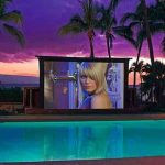 MovieMovil, la empresa de cine al aire libre, ofrece noches cinematográficas únicas en entornos paradisíacos Proyección de cine al aire libre junto a una piscina en un entorno tropical, con una gran pantalla que muestra una película bajo un cielo de tonos púrpura al atardecer. El ambiente incluye palmeras y un espacio relajante. MovieMovil, la empresa de cine al aire libre